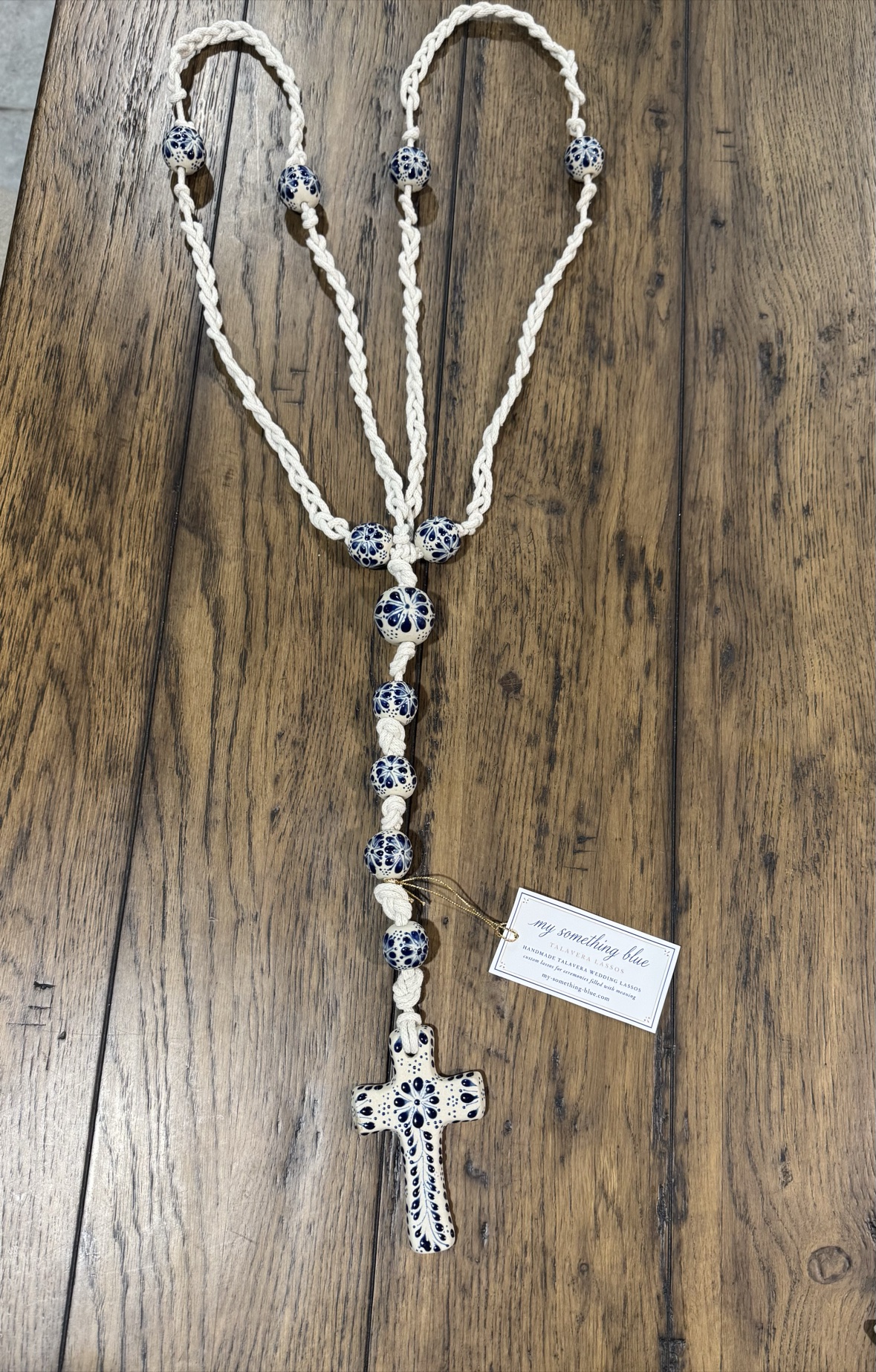 Ready-made Talavera lasso with braided cotton cord, blue-and-white beads, ceramic cross, and product tag on a wood surface