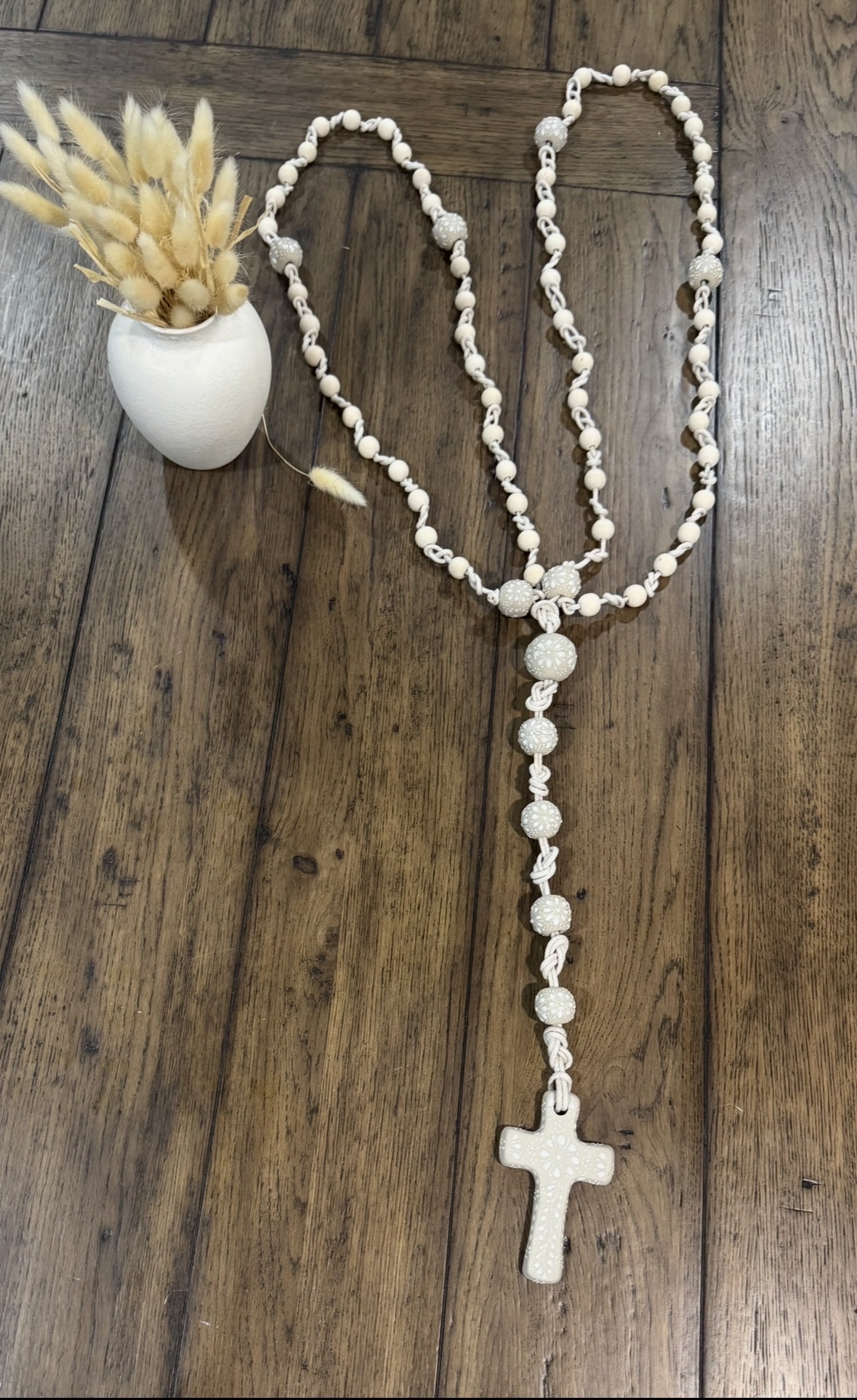 Ready-made ivory wedding lasso with natural wooden beads, ivory Talavera-style beads, a ceramic cross, and a small vase on a wood floor